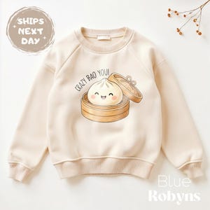 May include: Cream-colored sweatshirt with a crew neck and long sleeves. The front features a cartoon illustration of a smiling bao bun in a bamboo steamer, with the text "Crazy Bao You!". The brand name "Blue Robyn" is printed at the bottom right.
