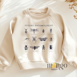 May include: A cream-colored sweatshirt with the text "FUTURE ENTOMOLOGIST" printed on the front. The text is surrounded by illustrations of various insects, including a dragonfly, ant, cricket, treehopper, mosquito, spider, moth, fly, bee, cicada, wasp, and beetle.