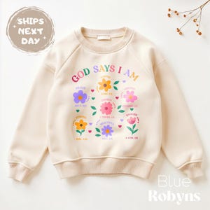 Op de afbeelding: Cr&egrave;mekleurige sweatshirt met de tekst "GOD SAYS I AM" en kleurrijke bloemengraphics. De sweatshirt heeft de woorden "Brave", "Worthy", "Chosen", "Beautiful" en "Enough". De merknaam "Blue Robyns" staat onderaan.