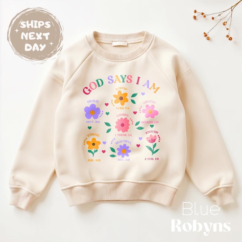 Op de afbeelding: Cr&egrave;mekleurige sweatshirt met de tekst "GOD SAYS I AM" en kleurrijke bloemengraphics. De sweatshirt heeft de woorden "Brave", "Worthy", "Chosen", "Beautiful" en "Enough". De merknaam "Blue Robyns" staat onderaan.