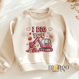 Peut inclure: Un sweat-shirt crème avec une pelleteuse rose qui creuse un tas de cœurs roses. Le texte "I DIG YOU" est imprimé en lettres roses au-dessus de la pelleteuse.