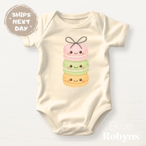 May include: A cream-colored baby onesie with a stack of three macarons with smiling faces. The macarons are pink, green, and yellow.
