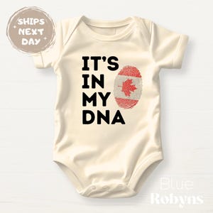 May include: A cream-coloured baby bodysuit featuring the text "IT'S IN MY DNA" in black. The design includes a fingerprint of the Canadian flag. The bodysuit has short sleeves and a round neckline.