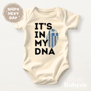 May include: Cream-colored baby bodysuit with the text "IT'S IN MY DNA" in black. The "A" in DNA is replaced with a blue and white fingerprint design resembling the Greek flag. A round sticker on the top left says "SHIPS NEXT DAY".
