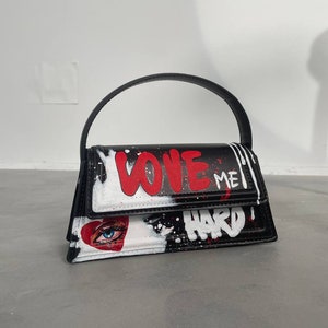 Può includere: Borsa a mano nera e bianca con un cuore rosso e un'immagine di un occhio. La borsa ha un manico nero e le parole "LOVE ME HARD" scritte in vernice rossa e bianca.