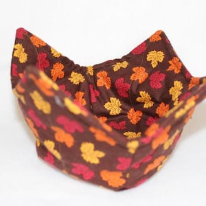 May include: A fabric bowl cozy featuring a dark brown background with a repeating pattern of small autumn leaves in shades of red, orange, and yellow. The bowl cozy has four pointed corners and is constructed with visible stitching throughout the fabric.