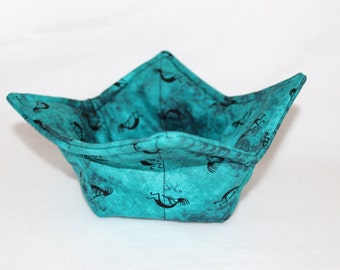 May include: A teal blue fabric bowl with a black and white pattern of stylised figures. The bowl is folded and has a square shape.