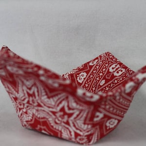 Peut inclure: Un chauffe-bol en tissu arborant un motif bandana rouge et blanc vif avec des dessins géométriques et floraux. Ce support de forme carrée présente quatre coins repliés et est fabriqué en textile matelassé avec des coutures visibles.