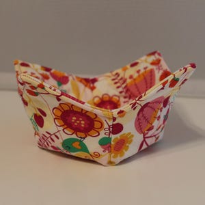 Puede incluir: Un cuenco de tela blanco con un patrón floral en tonos rosa, naranja y amarillo. El cuenco está doblado en forma de cuenco y es perfecto para guardar objetos pequeños.