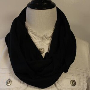 May include: A black infinity scarf made of a soft, stretchy fabric. The scarf is worn around the neck and drapes down the front of the chest.