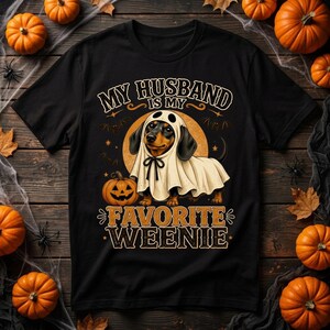 Puede incluir: Una camiseta negra con el gráfico de un perro salchicha vestido con un disfraz de fantasma blanco con un lazo negro. El diseño incluye una calabaza, murciélagos y el texto "MY HUSBAND IS MY FAVORITE WEENIE" en letras naranjas y blancas.