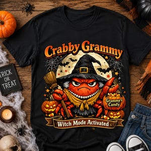 Op de afbeelding: Een zwart t-shirt met een afbeelding van een oranje krab met een heksenhoed, een gele sjaal en een parelketting. De krab houdt een bezem en een pompoenemmer vast met het label Grammy's Candy. Tekst Crabby Grammy en Witch Mode Activated.