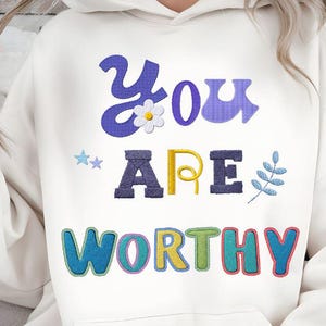 May include: Cream-coloured hoodie featuring the embroidered phrase "You Are Worthy" in vibrant, playful lettering. The design incorporates a daisy, stars, and a sprig of leaves. A comfortable and stylish garment.