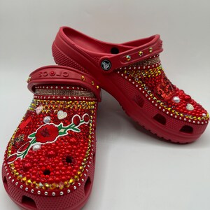 K&ouml;nnte beinhalten: Rote Crocs Clogs, reich verziert mit roten, goldenen und klaren Strasssteinen, Perlen und einem floralen Design. Auf einem Clog ist das Wort "LOVE" zu sehen. Die Clogs sind mit einer Vielzahl von glitzernden Edelsteinen verziert.