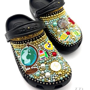 May include: Black Crocs decorated with gold and blue rhinestones, pearls, and various school-themed charms, including a globe, books, and a school bus. The charms are arranged in a whimsical pattern on the shoes. The text "Let's Study!" is visible on one of the charms.