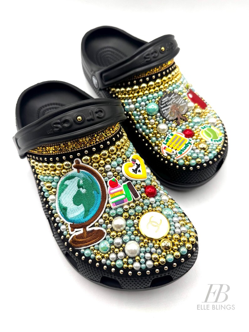 May include: Black Crocs decorated with gold and blue rhinestones, pearls, and various school-themed charms, including a globe, books, and a school bus. The charms are arranged in a whimsical pattern on the shoes. The text "Let's Study!" is visible on one of the charms.