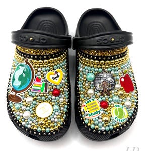 May include: Black and gold custom decorated clogs with a school theme. The clogs are covered in rhinestones, pearls, and charms. The charms include a globe, books, an apple, a heart, and a tennis ball. The text "Let's Study!" and "Hello School" are also included.