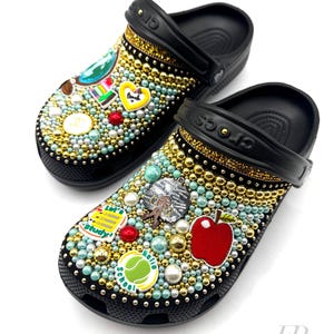 May include: Black and white Crocs decorated with colourful school-themed charms, including an apple, a tennis ball, and a book. The charms are surrounded by gold and white beads.