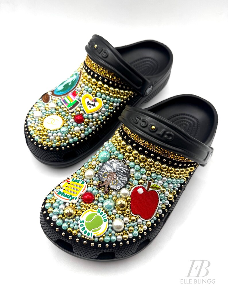 May include: Black and white Crocs decorated with colourful school-themed charms, including an apple, a tennis ball, and a book. The charms are surrounded by gold and white beads.