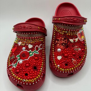 K&ouml;nnte beinhalten: Ein Paar rote Crocs-Sandalen, reich verziert mit roten Perlen, Strasssteinen und Charms. Die Sandalen haben ein Blumendesign, Herzen und das Wort "LOVE". Die R&auml;nder sind mit goldfarbenen Strasssteinen besetzt.