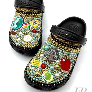 May include: Black Crocs decorated with rhinestones, pearls, and colourful school-themed charms, including a globe, an apple, a stack of books, and a tennis ball with "Hello School" written on it.