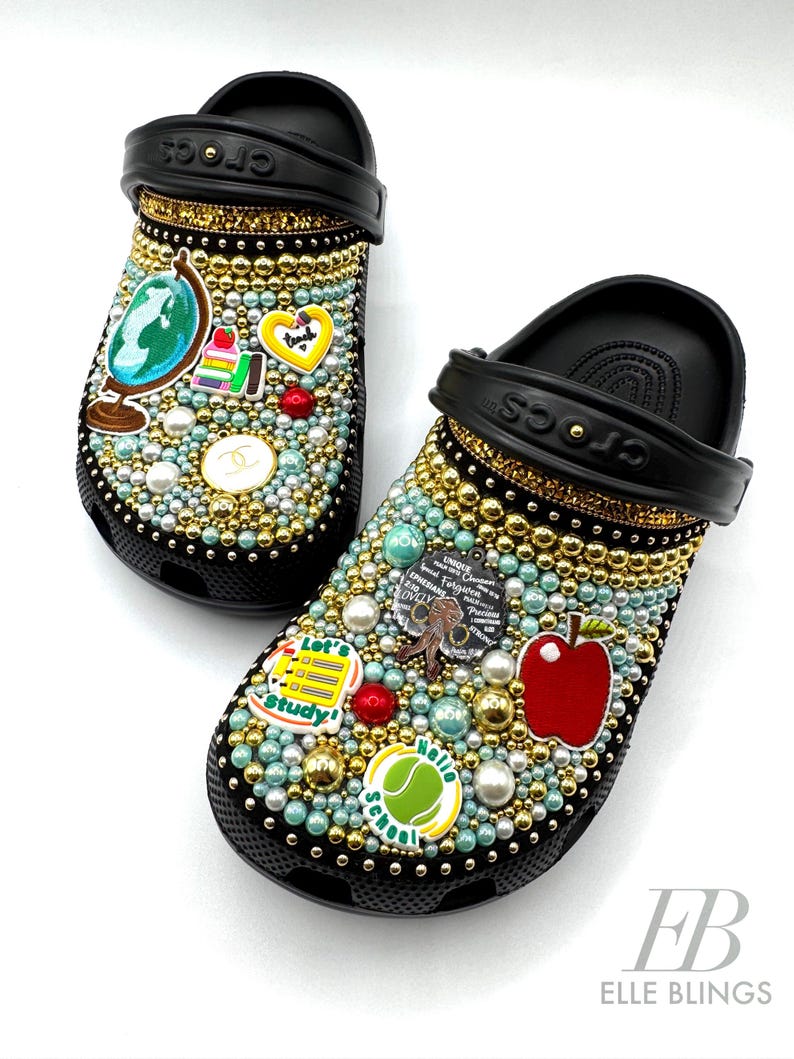 May include: Black Crocs decorated with rhinestones, pearls, and colourful school-themed charms, including a globe, an apple, a stack of books, and a tennis ball with "Hello School" written on it.