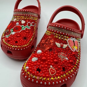 K&ouml;nnte beinhalten: Rote Clogs, reich verziert mit roten Perlen, Strasssteinen und dekorativen Anh&auml;ngern. Einer der Clogs hat ein Blumendesign, der andere ein paillettenbesetztes Herz und Schmetterlingsakzente. Die Clogs haben einen Riemen.