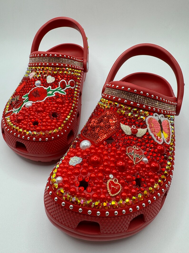 K&ouml;nnte beinhalten: Rote Clogs, reich verziert mit roten Perlen, Strasssteinen und dekorativen Anh&auml;ngern. Einer der Clogs hat ein Blumendesign, der andere ein paillettenbesetztes Herz und Schmetterlingsakzente. Die Clogs haben einen Riemen.