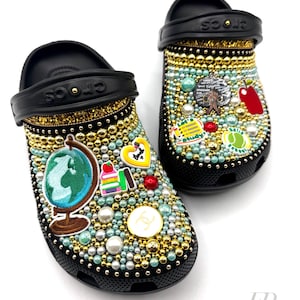 May include: A pair of black Crocs decorated with colourful rhinestones and charms. The charms include a globe, books, an apple, a tennis ball, and a heart. The Crocs are also decorated with gold glitter and pearls.