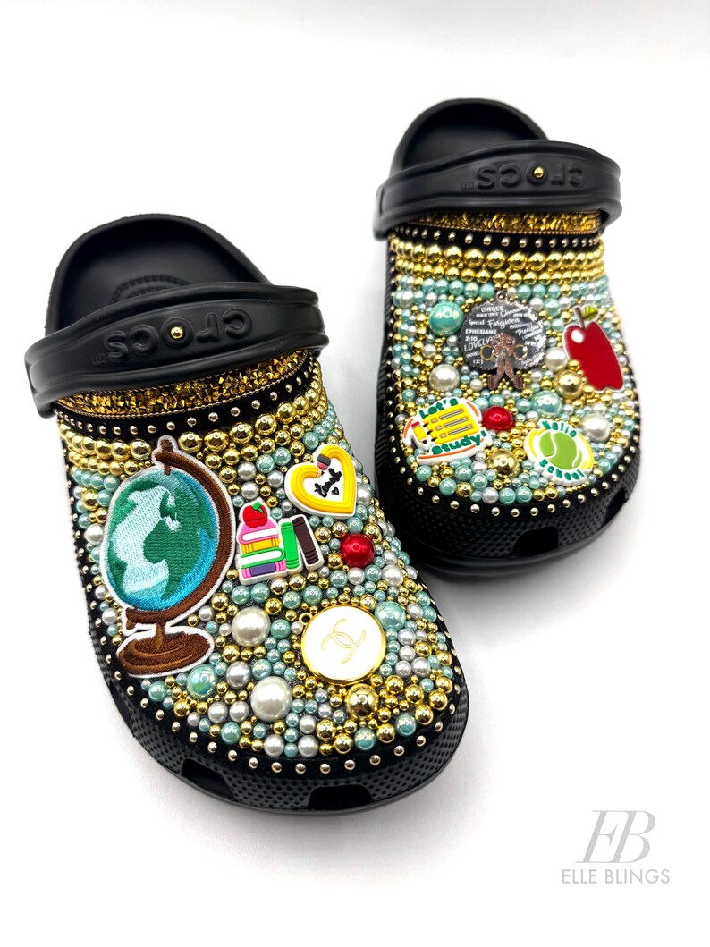 May include: A pair of black Crocs decorated with colourful rhinestones and charms. The charms include a globe, books, an apple, a tennis ball, and a heart. The Crocs are also decorated with gold glitter and pearls.