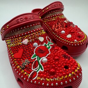 K&ouml;nnte beinhalten: Ein Paar rote Crocs-Schuhe, reich verziert mit roten und goldenen Strasssteinen, Perlen und gestickten Blumen- und Herzmotiven. Das Wort "LOVE" ist auf einem Schuh zu sehen.