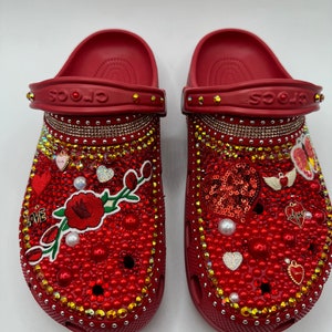 K&ouml;nnte beinhalten: Ein Paar rote Crocs-Sandalen, reich verziert mit roten und goldenen Strasssteinen, Perlen und dekorativen Anh&auml;ngern. Die Sandalen zeigen ein Blumendesign, Herzen und das Wort "LOVE".