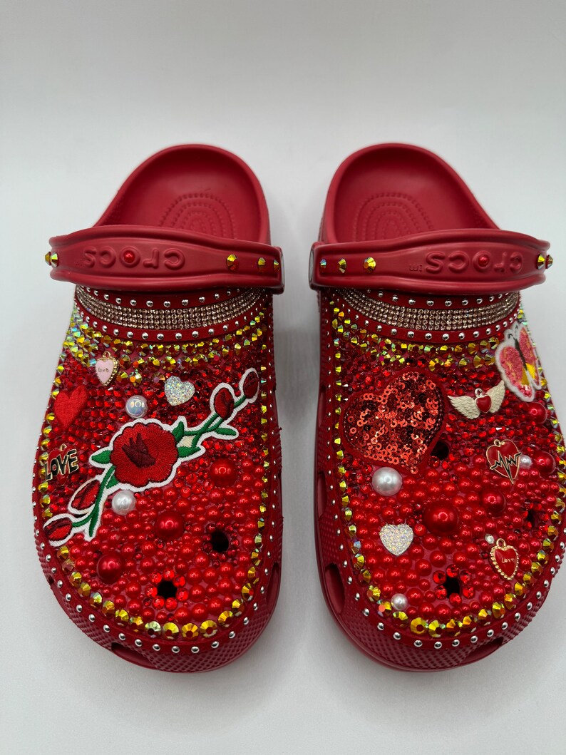 K&ouml;nnte beinhalten: Ein Paar rote Crocs-Sandalen, reich verziert mit roten und goldenen Strasssteinen, Perlen und dekorativen Anh&auml;ngern. Die Sandalen zeigen ein Blumendesign, Herzen und das Wort "LOVE".