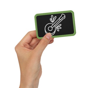 May include: A green and black embroidered patch with a white outline of a banjo. The patch is held in a hand.