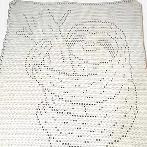 May include: Cream-colored crochet blanket featuring a detailed design of a face with a floral crown. The blanket is rectangular and has a textured appearance due to the crochet stitches. The design is created with small, dark-colored stitches.