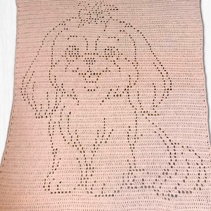 May include: A pale pink crocheted blanket with a detailed outline of a dog. The dog's face, ears, and body are clearly defined within the crocheted pattern. The blanket is rectangular and appears to be handmade.