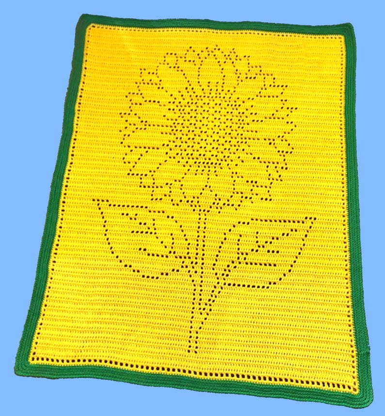 Puede incluir: Una manta rectangular de ganchillo que presenta un campo amarillo brillante con un motivo central de girasol creado mediante puntos calados. La manta est&aacute; rematada con un borde verde s&oacute;lido que enmarca toda la pieza rectangular amarilla.