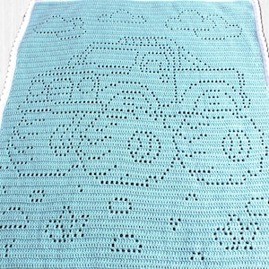 May include: A light blue crochet blanket featuring a central design of a tractor with wheels and a cab, surrounded by small clouds and circular shapes. The rectangular blanket is finished with a white scalloped border along the edges.