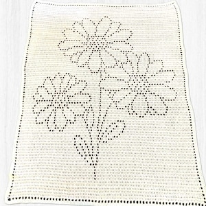 May include: A white crocheted blanket featuring a floral design. The blanket has a rectangular shape with a black dotted border. The floral pattern includes three large flowers with detailed petals and stems.