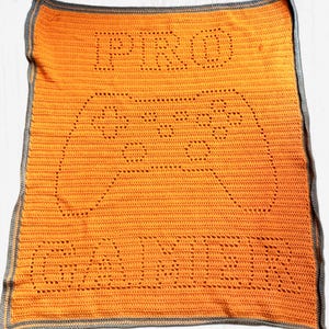 May include: A rectangular orange crochet blanket featuring a dark gray border. The center displays a large outline of a video game controller created with open stitches. The text PRO is positioned above the controller and GAMER below it.