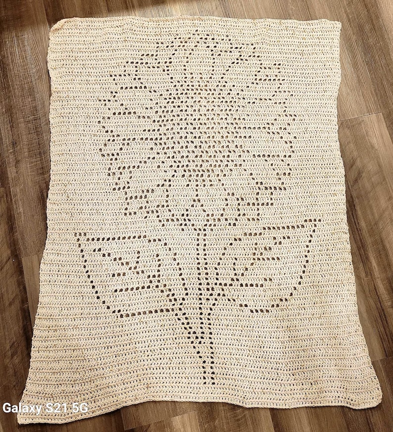 Puede incluir: Una manta rectangular de ganchillo en color blanco roto que presenta un gran motivo de girasol centrado, creado con puntos calados. El dise&ntilde;o muestra una cabeza de flor detallada con p&eacute;talos radiantes y un tallo con dos hojas.