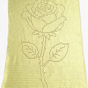 May include: A rectangular pale yellow crochet blanket featuring a central rose motif created with openwork stitches. The design displays a single rose bloom with a stem and two leaves, formed by the negative space within the solid crochet fabric.