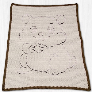 May include: A cream-colored crocheted blanket featuring a brown outline of a bear holding a small object. The blanket has a brown border. The bear's eyes and nose are detailed with small, evenly spaced holes.