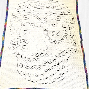 May include: A rectangular crochet blanket featuring a cream-colored base with a large, intricate black skull design created through open-work stitching. The blanket is finished with a vibrant, multi-colored rainbow yarn border in a scalloped pattern.