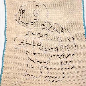 May include: A rectangular crochet blanket featuring a cream-coloured base with a dark brown outline of a turtle character. The blanket is finished with a bright turquoise blue border along all four edges, created with a consistent stitch pattern.