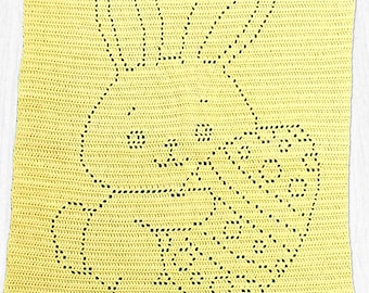 Pode incluir: Uma manta retangular de crochê amarela com um design central de um coelho segurando um ovo estampado. O design é criado usando pontos abertos contra um fundo sólido. O texto HAPPY EASTER está feito em crochê na borda inferior.
