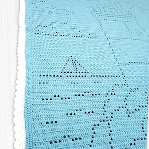 May include: A light blue crochet blanket featuring a lighthouse, a sailboat, and cloud motifs created with openwork stitches. The blanket is finished with a white scalloped border along the edge, showcasing a detailed nautical-themed pattern.