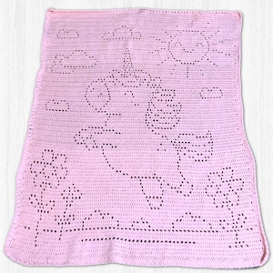 Peut inclure: Une couverture rectangulaire en crochet rose clair présentant un motif central de licorne. Le design inclut un soleil, des nuages et des fleurs créés par des points ajourés. La texture est uniforme sur toute la pièce, révélant un motif détaillé.