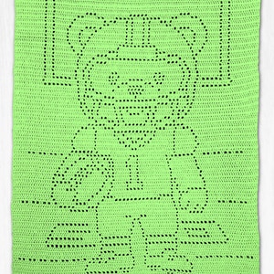 May include: A rectangular lime green crochet blanket featuring a central bear motif created with openwork stitches. The bear design is centred on the fabric, with the bear's head, torso, and limbs clearly defined by the negative space of the yarn.