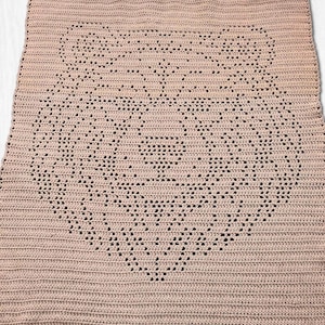 Pode incluir: Uma manta retangular de croch&ecirc; em uma cor rosa antigo s&oacute;lida. O tecido apresenta um design central de rosto de urso criado com uma t&eacute;cnica de croch&ecirc; fil&eacute;, onde espa&ccedil;os abertos no padr&atilde;o formam as orelhas, os olhos e o focinho do urso.